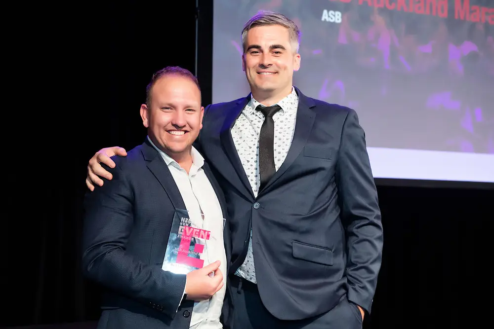 Two men in suits smiling on stage, one holding a NZEA Event Award trophy